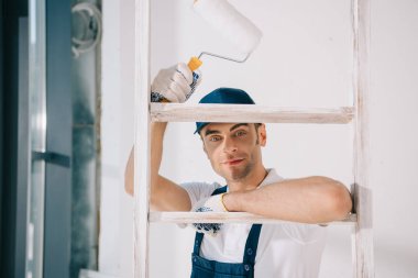 handsome young painter in uniform standing near ladder, holding paint roller and smiling at camera clipart