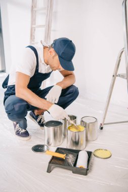 young painter in overalls and cap taking yellow paint from can with paintbrush clipart