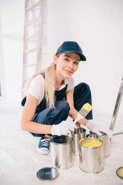 beautiful young painter in overalls and cap holding paintbrush near can with yellow paint clipart