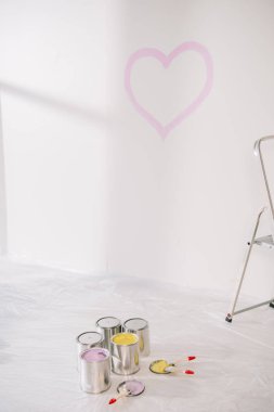 Beyaz duvarda pembe kalbi olan bir oda, beyaz zeminde boya ve boya fırçaları olan teneke kutular, selofan kaplı.
