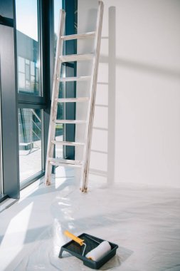 Geniş pencereli bir oda, beyaz duvarın yanındaki merdiven, yerde selofan kaplı tekerlekli tepside boya silindiri.