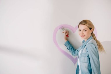 Beyaz duvarda fırçayla pembe kalbi boyayan mutlu genç bir kadın.