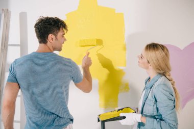 young woman holding roller tray while boyfriend painting wall in yellow with paint roller clipart