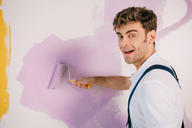 handsome young painter painting wall in pink color with paint roller and smiling at camera clipart