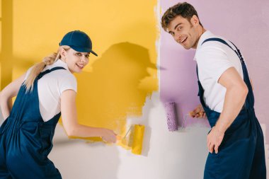 two cheerful painters looking at camera while painting wall in yellow and pink clipart