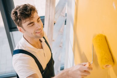 handsome young painter smiling while painting wall in yellow color with paint roller clipart