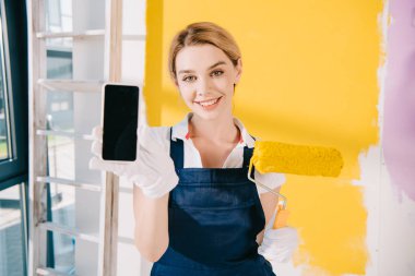 Üniformalı güzel bir ressam boya silindiri tutarken boş ekranlı akıllı telefonu gösteriyor.