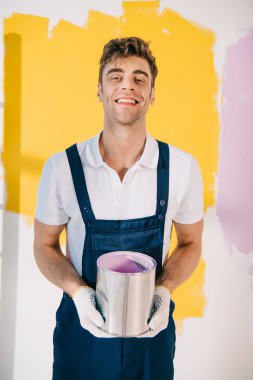 cheerful painter in uniform holding can with paint and smiling at camera clipart