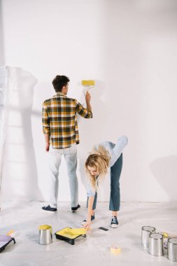 young woman putting paint roller into roller tray with yellow paint while boyfriend painting wall clipart