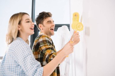 happy young couple painting white wall in yellow with paint rollers clipart