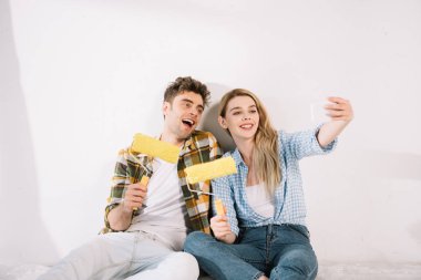 Beyaz duvarın yanında otururken ve sarı boya rulosu tutarken selfie çeken neşeli genç çift.