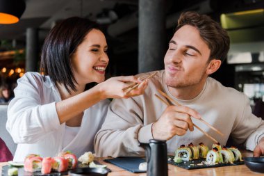 Lokantada neşeli bir adamın yanında lezzetli suşiyle yemek çubuğu tutan mutlu kadının seçici odağı. 
