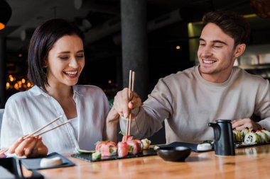 Lokantada yakışıklı bir adamın yanında lezzetli suşiyle yemek çubuğu tutan mutlu kadının seçici odağı. 