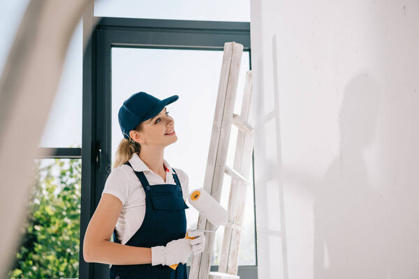 selective focus of young painter in uniform standing near ladder, holding paint roller and looking at white wall