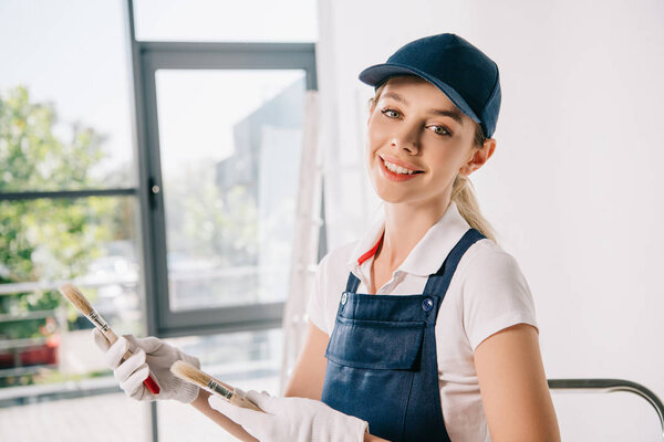 attractive young painter in uniform holding paintbrushes and smiling at camera