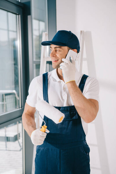 handsome young painter in uniform looking away while holding paint roller and talking on smartphone