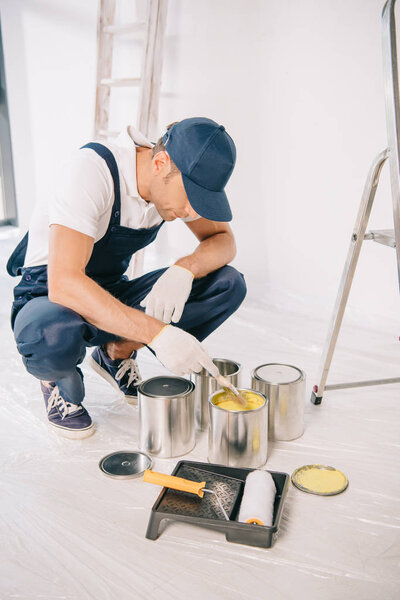 young painter in overalls and cap taking yellow paint from can with paintbrush