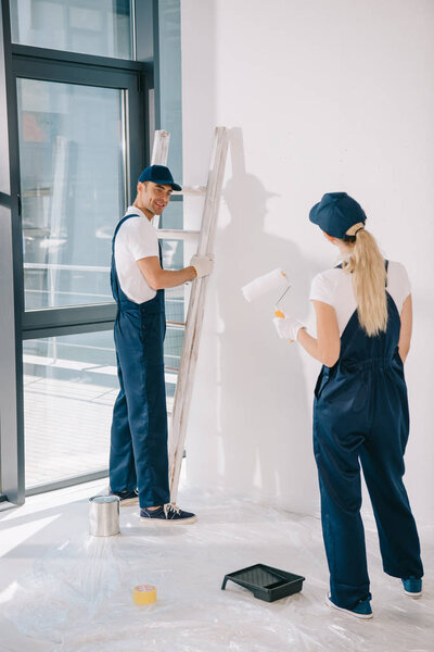 handsome painter standing near ladder while pretty colleague holding paint roller