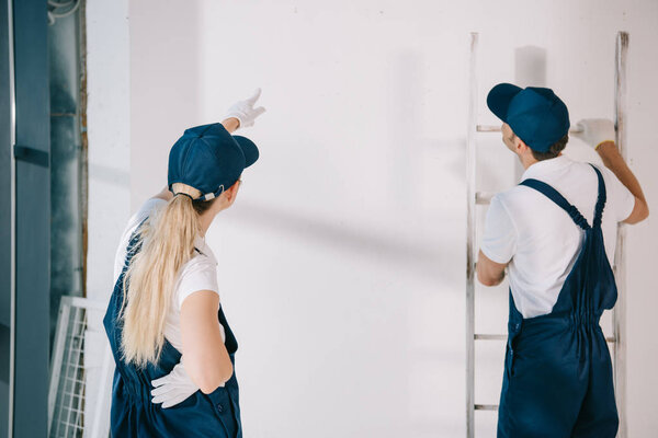 young painter in uniform pointing with finger at white wall while standing near coworker holding ladder