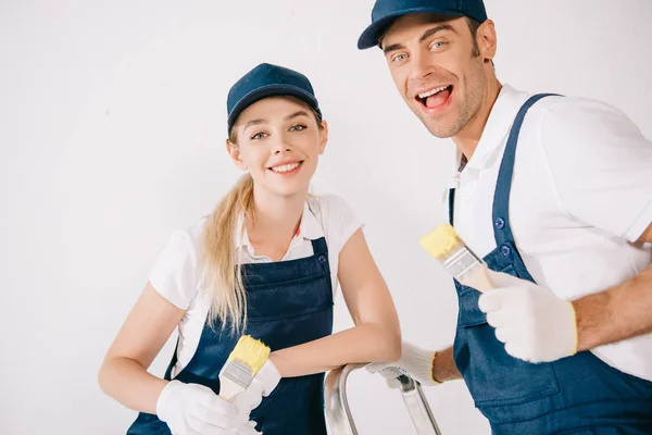 Dos Pintores Alegres Uniforme Sosteniendo Pinceles Sonriendo Cámara —  Fotos de Stock