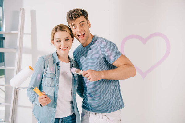 happy young couple looking at camera while holding pink paintbrush and paint roller