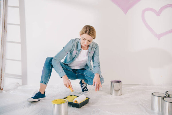 pretty young woman putting paint roller into roller tray with yellow paint