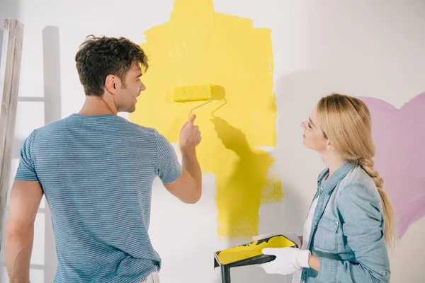 Young Woman Holding Roller Tray While Boyfriend Painting Wall Yellow — Stock Photo, Image