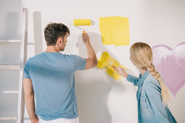 young couple painting white wall in yellow with paint rollers