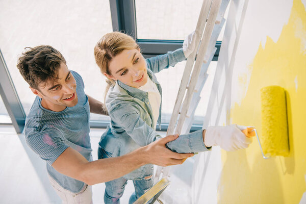 high angle view of handsome young man helping girlfriend painting wall in yellow