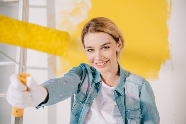 selective focus of pretty young woman holding yellow paint roller and smiling at camera