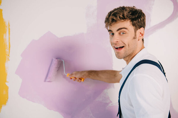 handsome young painter painting wall in pink color with paint roller and smiling at camera