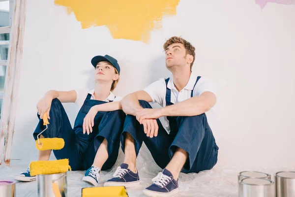 exhausted painters in overalls sitting on floor near wall