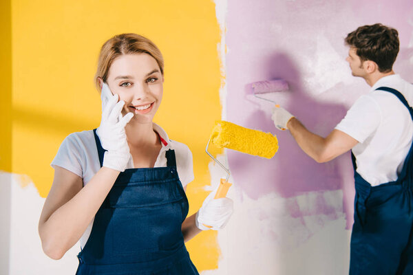 attractive painter in uniform talking on smartphone while handsome colleague painting wall