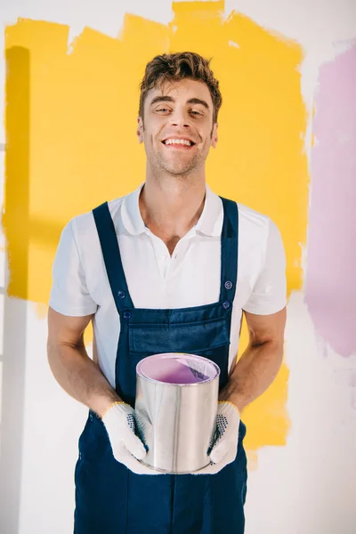 cheerful painter in uniform holding can with paint and smiling at camera