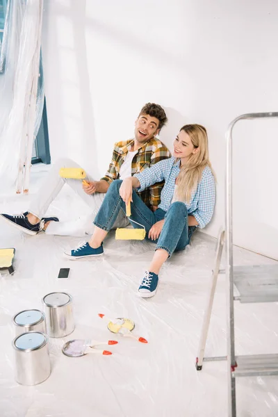 快乐的年轻夫妇坐在白墙边 手里拿着黄色的油漆滚筒 — 图库照片