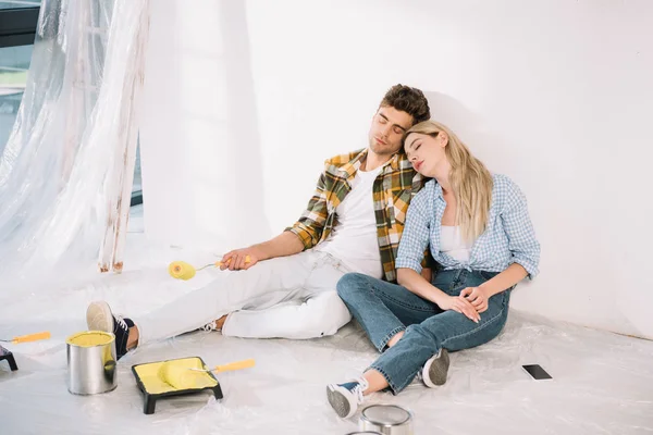 exhausted couple sleeping while sitting by white wall near roller tray with yellow paint