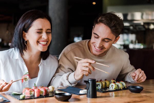 счастливый мужчина и веселая женщина смеются, держа палочки с вкусными суши в ресторане
 