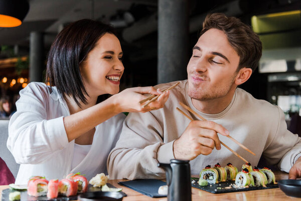 selective focus of happy woman holding chopsticks with tasty sushi near cheerful man in restaurant 