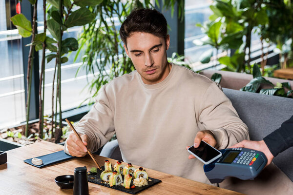 cropped view of waitress holding payment terminal near handsome man with smartphone 