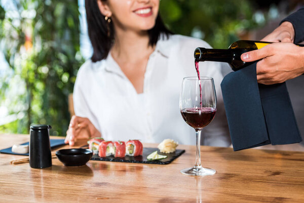 обрезанный вид официанта наливая красное вино в бокал рядом счастливая женщина
 