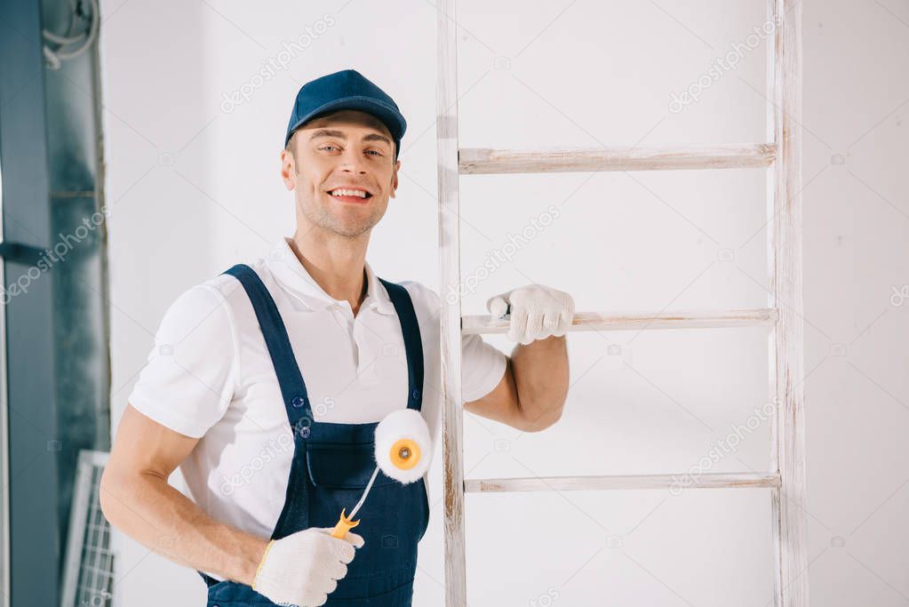 handsome young painter in uniform smiling at camera while standing near ladder and holding paint roller