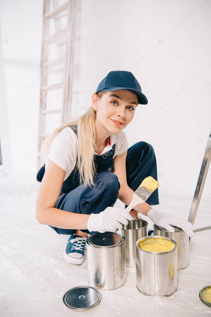 beautiful young painter in overalls and cap holding paintbrush near can with yellow paint