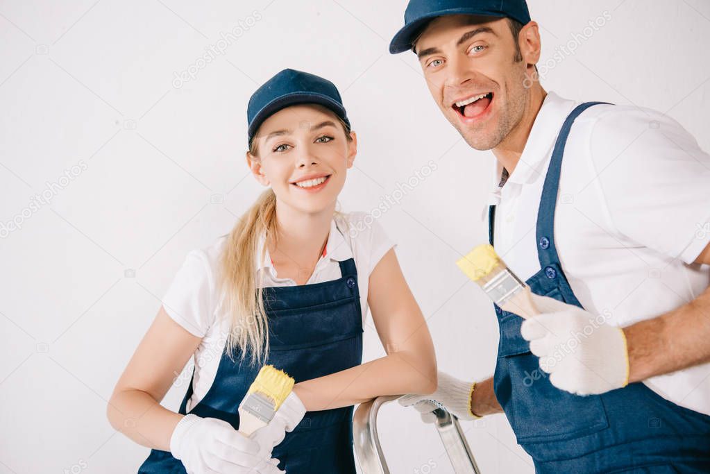 two cheerful painters in uniform holding paintbrushes and smiling at camera