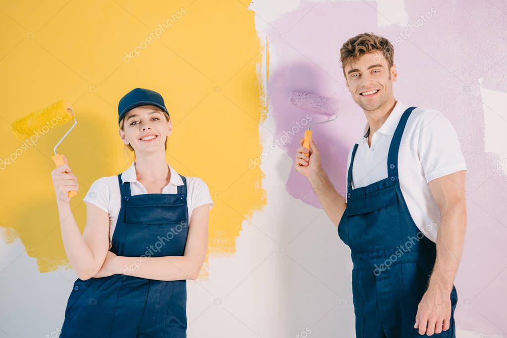 Young painters in overalls holding paint rollers and smiling at camera