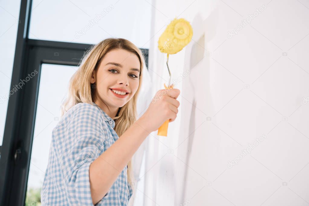 cheerful young woman holding yellow paint roller while standing near white wall