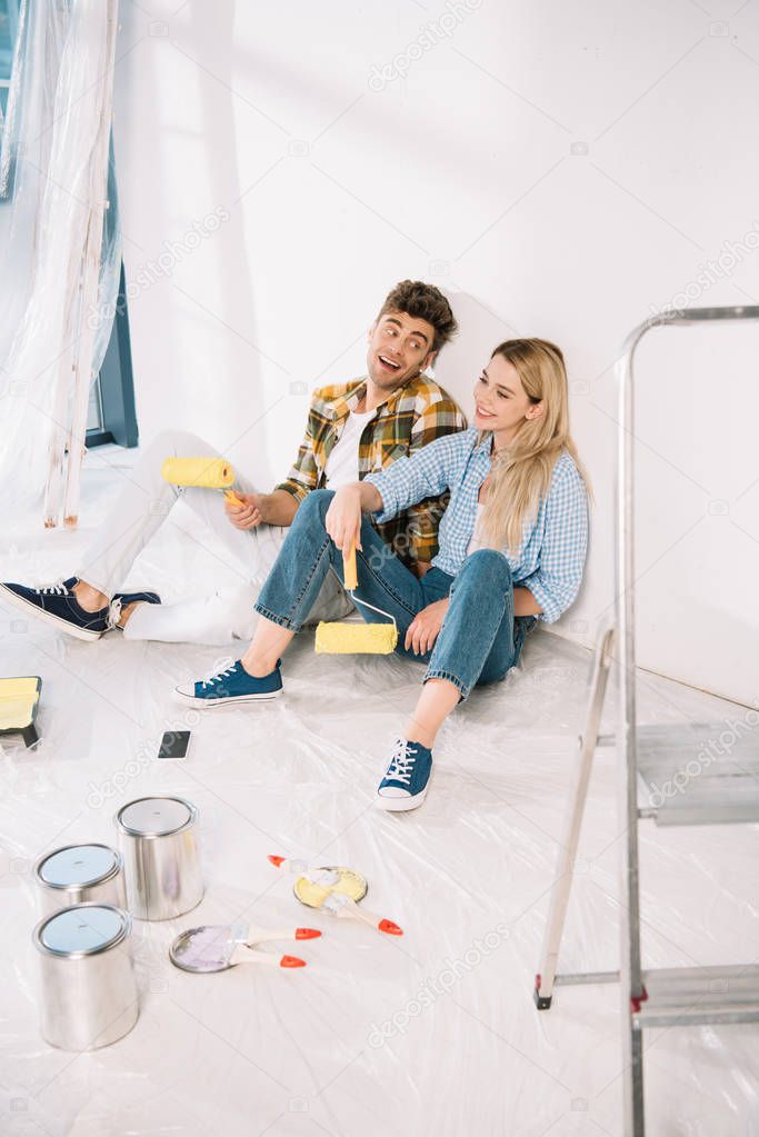 cheerful young couple sitting near white wall and holding yellow paint rollers