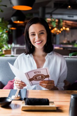 selective focus of cheerful woman sitting in restaurant and holding menu  clipart