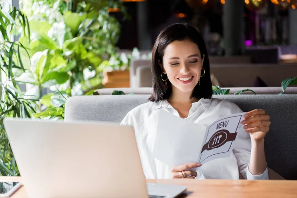 happy woman holding menu near laptop in restaurant