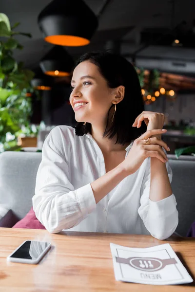 快乐的年轻女性坐在靠近菜单和智能手机的地方 桌上有空白的屏幕 — 图库照片
