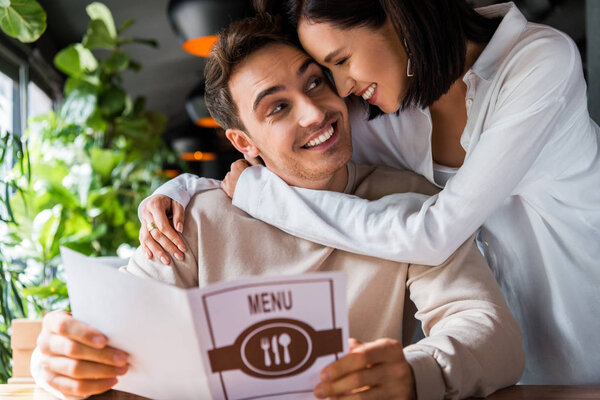 happy woman hugging cheerful man with menu in restaurant 